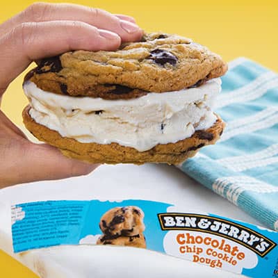 Hand holds a Ben & Jerry's ice cream sandwich. In the background a cutting board is visible. The background is yellow.